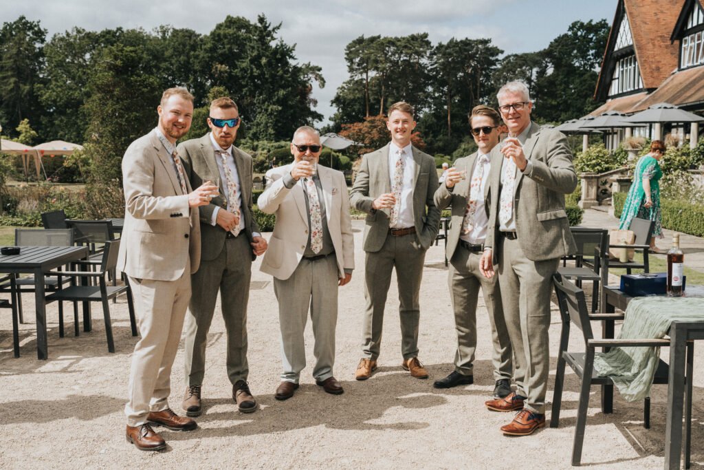 Six men in light suits and floral ties stand outdoors, smiling and holding drinks. They are gathered on a patio with empty tables and chairs, with greenery and a large building in the background on a sunny day. © Aimee Lince Photography, Wedding photographer in Lincolnshire