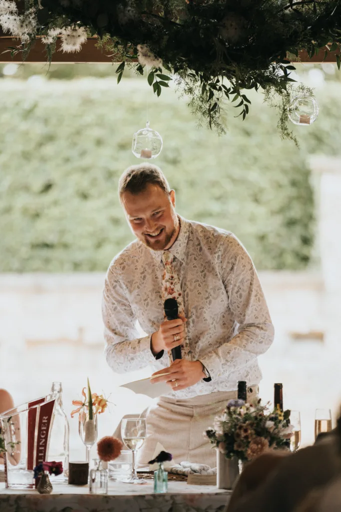 A smiling man in a patterned shirt stands behind a decorated table, holding a microphone as he prepares to speak at an outdoor event, possibly a wedding or celebration, with greenery and hanging glass ornaments above him. © Aimee Lince Photography, Wedding photographer in Lincolnshire