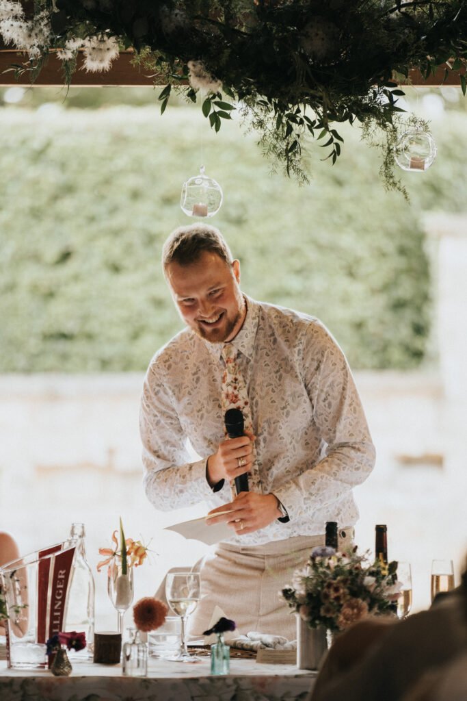 A smiling man in a patterned shirt stands behind a decorated table, holding a microphone as he prepares to speak at an outdoor event, possibly a wedding or celebration, with greenery and hanging glass ornaments above him. © Aimee Lince Photography, Wedding photographer in Lincolnshire