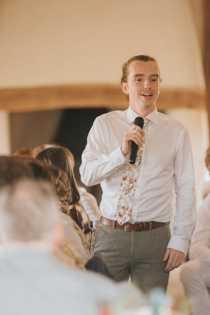 A young man in a white shirt and floral tie stands holding a microphone, speaking to a seated audience in a warmly lit indoor setting. © Aimee Lince Photography, Wedding photographer in Lincolnshire