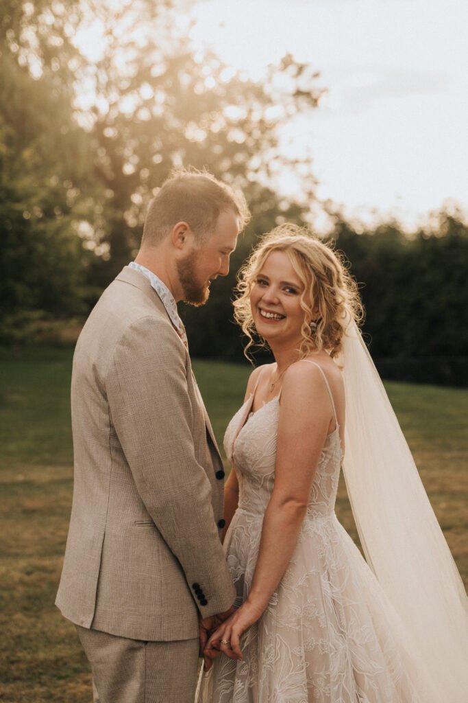 A bride in a white lace wedding dress and veil smiles, holding hands with a groom in a light beige suit, standing together outdoors with greenery and warm sunlight in the background. © Aimee Lince Photography, Wedding photographer in Lincolnshire