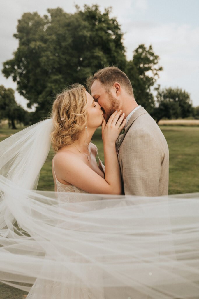 A bride and groom share a kiss outdoors. The bride's veil flows in the wind, wrapping around both of them. Trees and grass are visible in the background under a partly cloudy sky. © Aimee Lince Photography, Wedding photographer in Lincolnshire