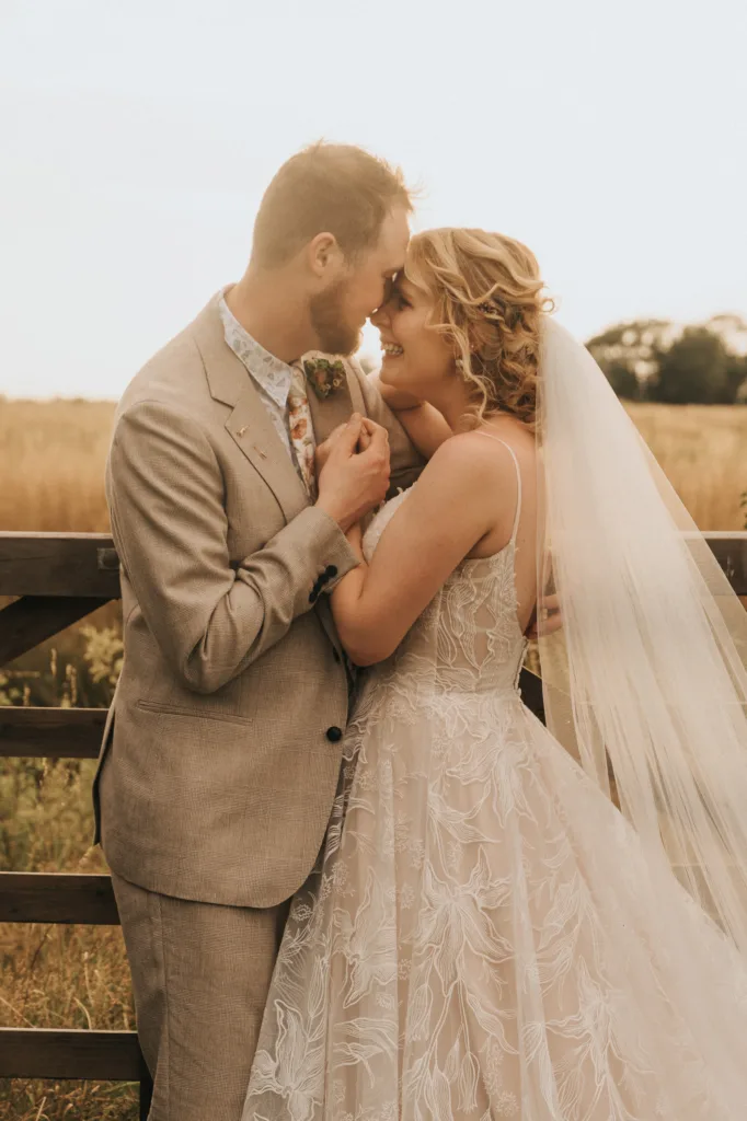 A bride and groom stand close together outdoors, smiling and touching foreheads. The bride wears a lace wedding dress and veil, while the groom wears a light suit. They are in front of a wooden fence with a field in the background. © Aimee Lince Photography, Wedding photographer in Lincolnshire
