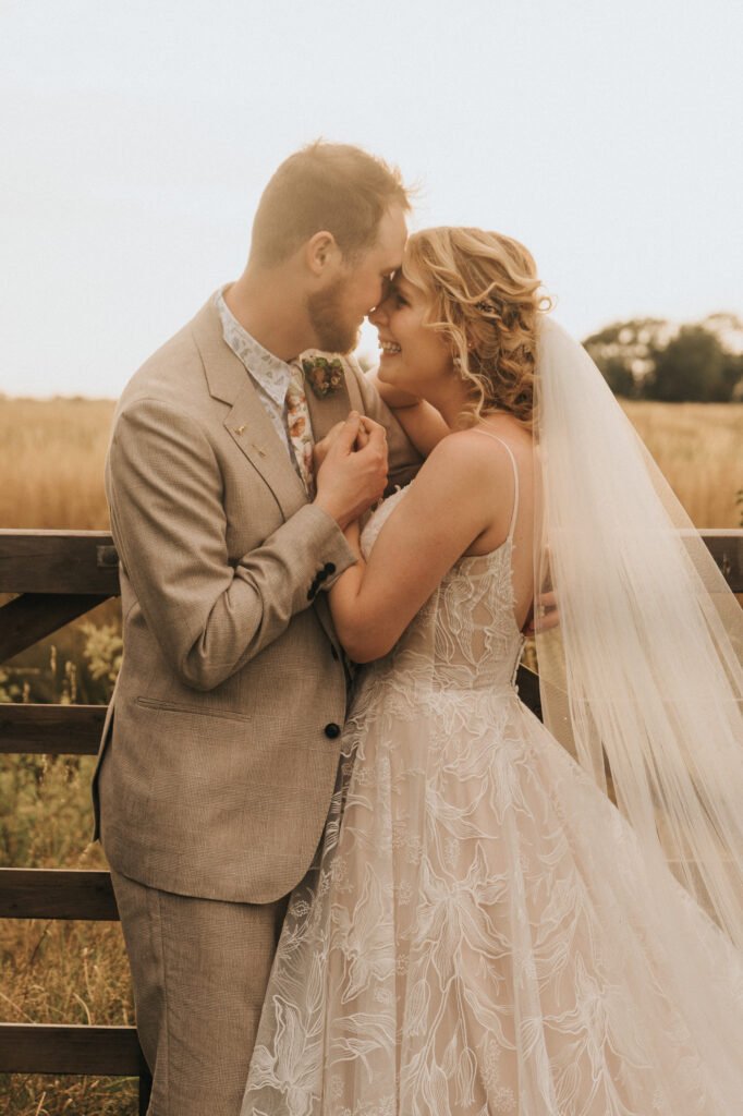 A bride and groom stand close together outdoors, smiling and touching foreheads. The bride wears a lace wedding dress and veil, while the groom wears a light suit. They are in front of a wooden fence with a field in the background. © Aimee Lince Photography, Wedding photographer in Lincolnshire