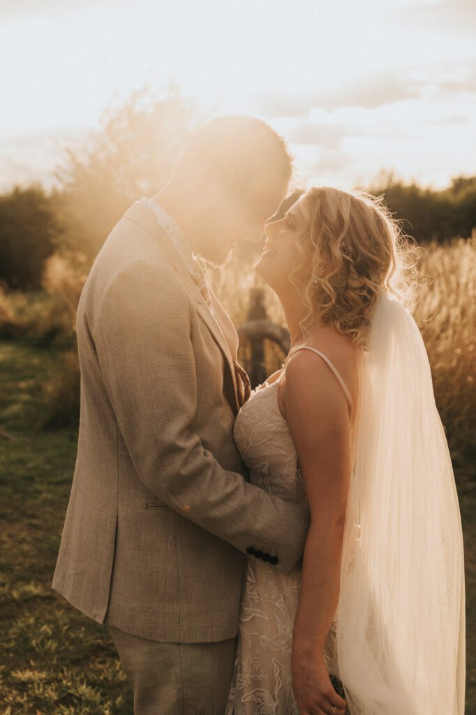 A bride and groom stand close together, smiling and touching foreheads outdoors at sunset. The bride wears a veil and lace dress, and the groom wears a light suit. Warm sunlight creates a romantic glow around them. © Aimee Lince Photography, Wedding photographer in Lincolnshire