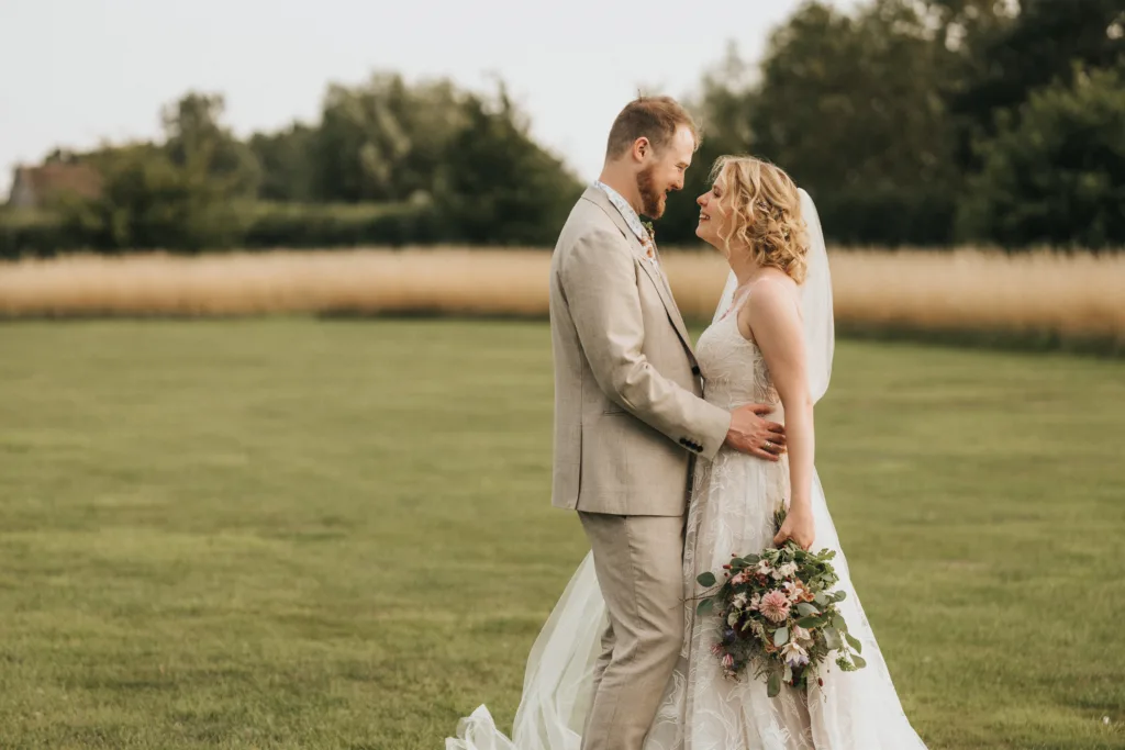 A bride and groom stand facing each other on a grassy field, smiling. The bride holds a bouquet of flowers and wears a white dress and veil, while the groom wears a light suit. Trees and tall grass are in the background. © Aimee Lince Photography, Wedding photographer in Lincolnshire