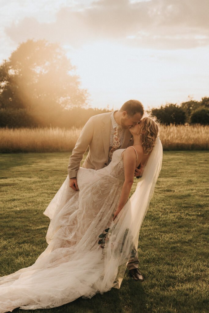 A bride and groom share a romantic kiss outdoors at sunset. The bride wears a flowing lace gown with a long veil, whilst the groom is dressed in a light suit. They stand on green grass with golden fields in the background. © Aimee Lince Photography, Wedding photographer in Lincolnshire