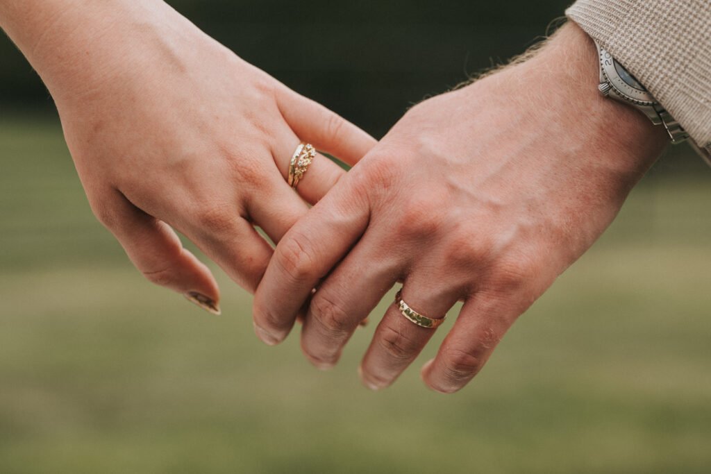 Two hands with wedding rings gently touching, set against a blurred outdoor background. The left hand has a gold ring with a detailed pattern; the right hand wears a simpler gold band and a watch. © Aimee Lince Photography, Wedding photographer in Lincolnshire