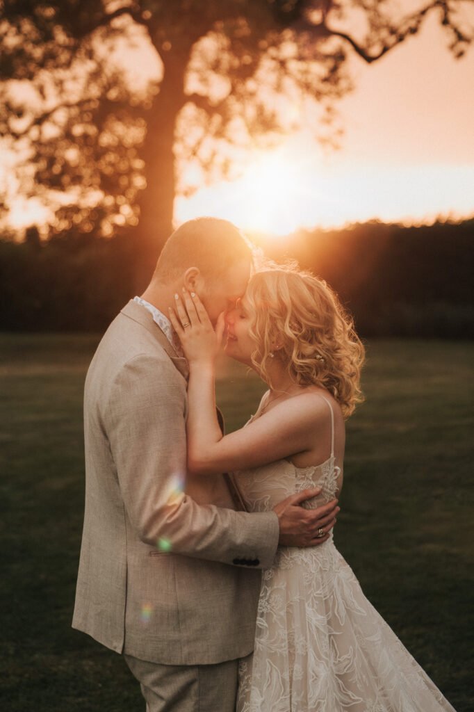 A bride and groom embrace closely at sunset in an outdoor setting, with warm sunlight glowing behind them and a tree silhouetted in the background. © Aimee Lince Photography, Wedding photographer in Lincolnshire