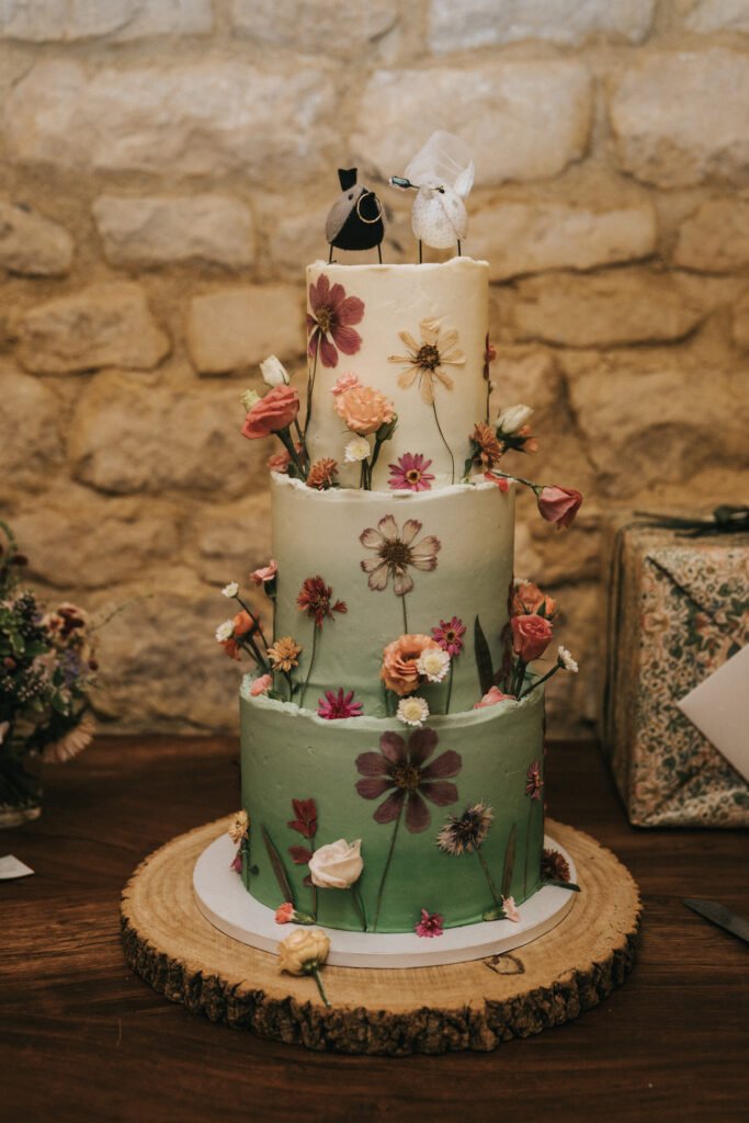 A three-tiered wedding cake decorated with pressed flowers and topped with bird figurines, sitting on a wooden slice, against a stone wall backdrop. © Aimee Lince Photography, Wedding photographer in Lincolnshire