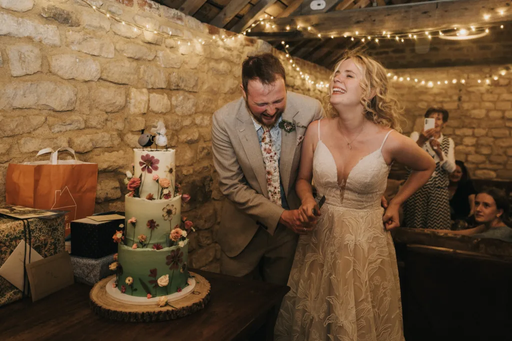 A bride and groom laugh joyfully as they cut a flower-decorated wedding cake together in a rustic venue decorated with fairy lights, surrounded by guests and presents. © Aimee Lince Photography, Wedding photographer in Lincolnshire