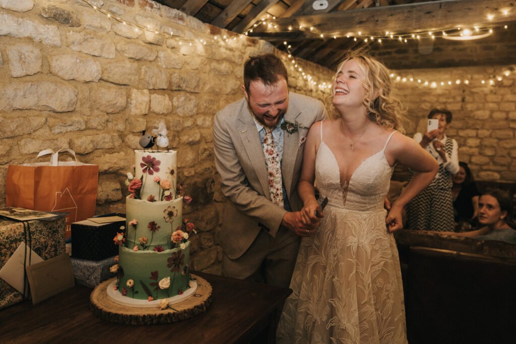 A bride and groom laugh joyfully as they cut a flower-decorated wedding cake together in a rustic venue decorated with fairy lights, surrounded by guests and presents. © Aimee Lince Photography, Wedding photographer in Lincolnshire