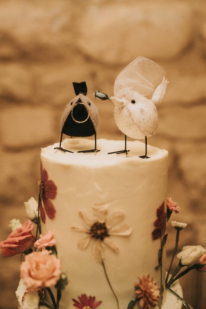 Two bird figurines, one with a black bow tie and top hat, the other with a white veil, sit atop a white cake decorated with flowers, resembling a wedding cake against a blurred beige background. © Aimee Lince Photography, Wedding photographer in Lincolnshire