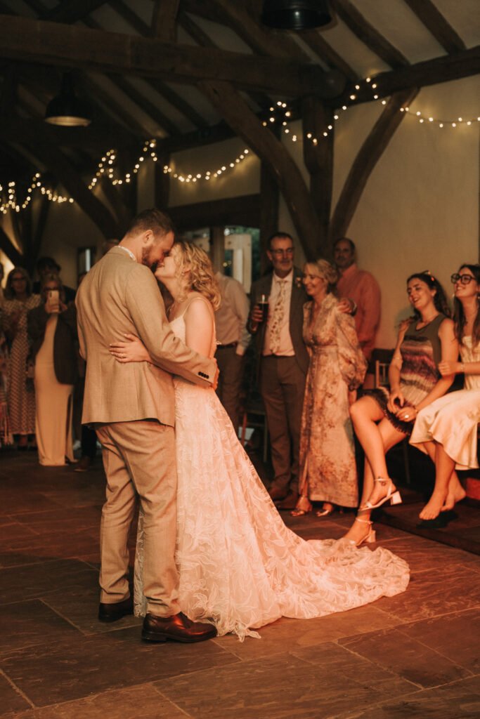 A bride and groom share a kiss while dancing at their wedding reception, surrounded by guests seated and standing, under warm fairy lights in a rustic wooden venue. © Aimee Lince Photography, Wedding photographer in Lincolnshire
