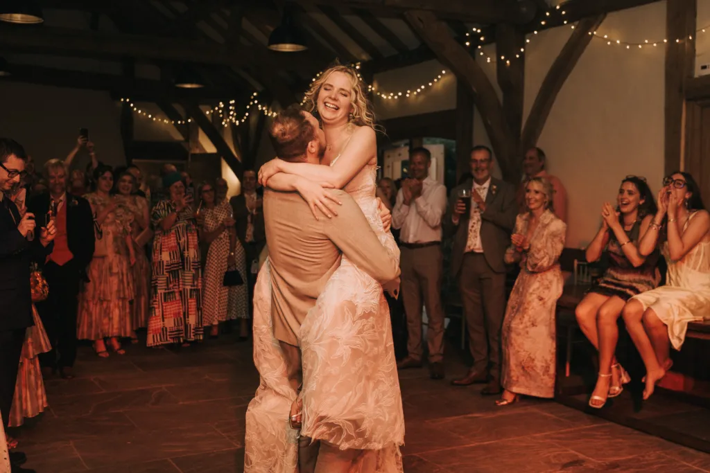 A bride and groom laugh and embrace whilst dancing at their wedding, surrounded by smiling guests. Warm lights and rustic wooden beams create a joyful, festive atmosphere in the reception venue. © Aimee Lince Photography, Wedding photographer in Lincolnshire