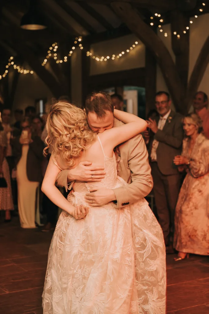 A bride and groom share a warm embrace whilst dancing at their wedding reception, surrounded by guests and soft fairy lights in a rustic, warmly lit venue. © Aimee Lince Photography, Wedding photographer in Lincolnshire