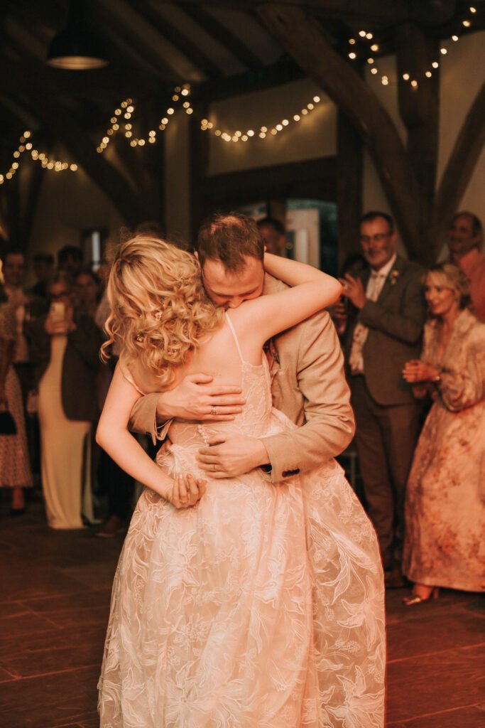 A bride and groom share a warm embrace whilst dancing at their wedding reception, surrounded by guests and soft fairy lights in a rustic, warmly lit venue. © Aimee Lince Photography, Wedding photographer in Lincolnshire