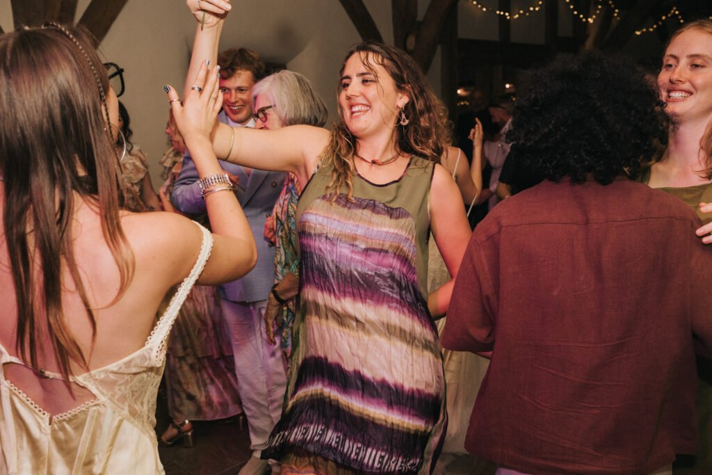A group of people, dressed in colourful and festive attire, dance together at a lively indoor event with fairy lights overhead. Everyone appears joyful and engaged in the celebration. © Aimee Lince Photography, Wedding photographer in Lincolnshire