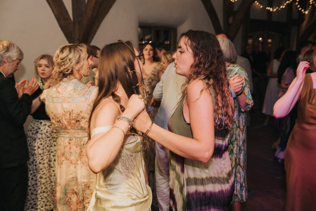 Two women are dancing closely together at a lively indoor event, surrounded by other people who are also dancing and socialising. The venue has wooden beams and fairy lights overhead. © Aimee Lince Photography, Wedding photographer in Lincolnshire