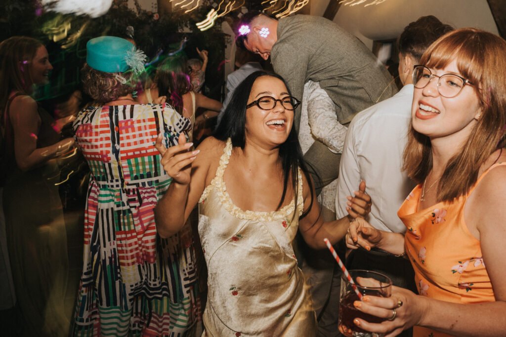 A group of people are joyfully dancing at a party. Two women in the foreground are smiling and holding drinks, surrounded by others in colourful outfits under festive lighting. The atmosphere is lively and celebratory. © Aimee Lince Photography, Wedding photographer in Lincolnshire