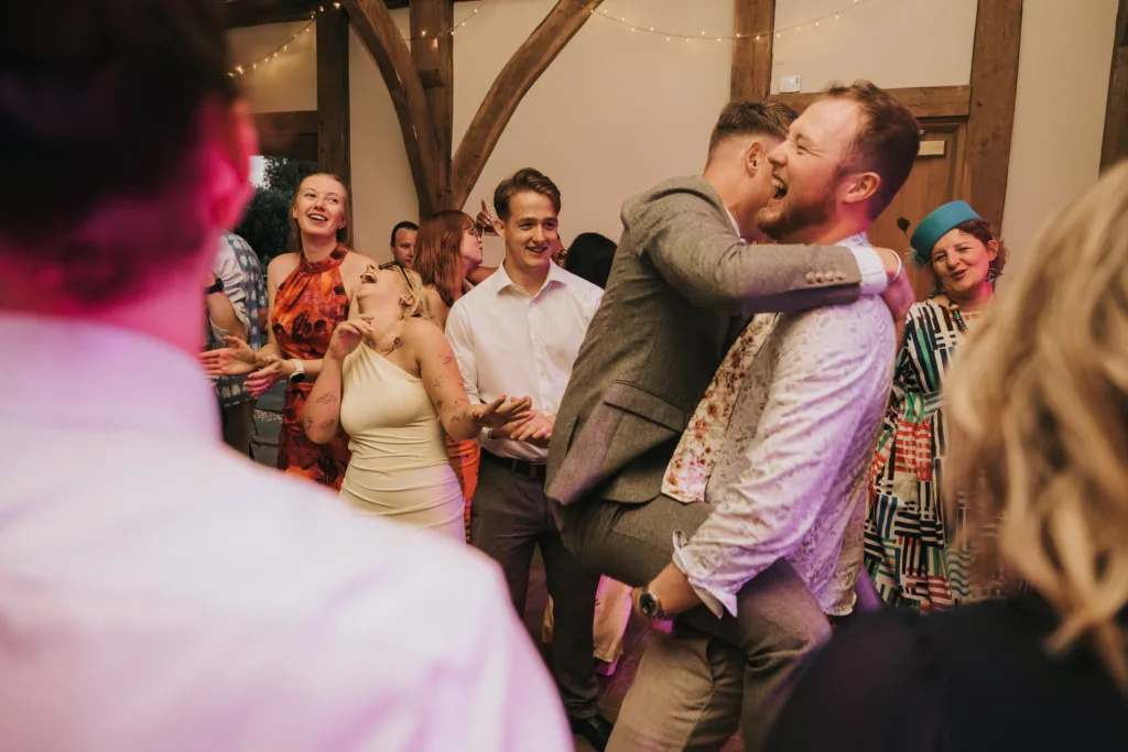 A lively group of people dance and laugh at a celebration. Two men in the centre joyfully embrace, one lifting the other off the ground. Others smile and cheer, creating a festive atmosphere in a decorated indoor venue. © Aimee Lince Photography, Wedding photographer in Lincolnshire