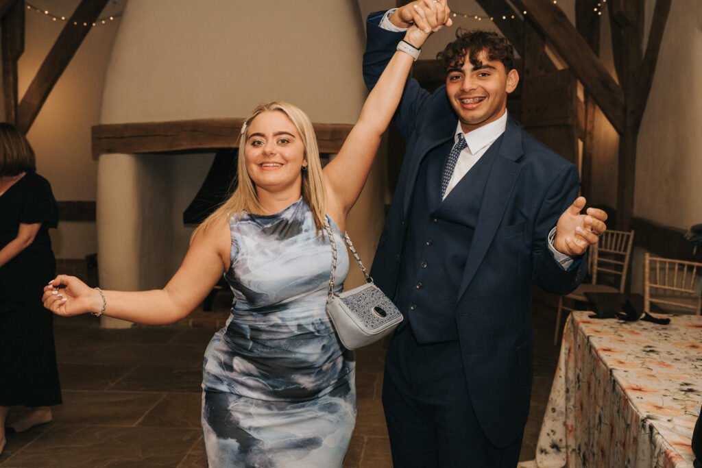 A smiling woman in a blue and white dress and a man in a blue suit are dancing together indoors, holding hands and appearing joyful. The setting features wooden beams and decorated tables in the background. © Aimee Lince Photography, Wedding photographer in Lincolnshire