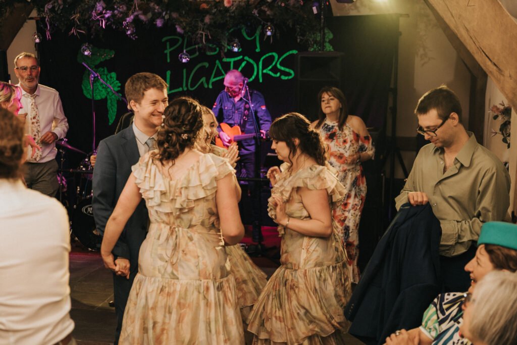 A lively group of people dance together at an indoor event, with three women in frilled dresses and a man in a suit in the centre. Musicians perform on a stage in the background under a sign that reads "Pussy Alligators. © Aimee Lince Photography, Wedding photographer in Lincolnshire