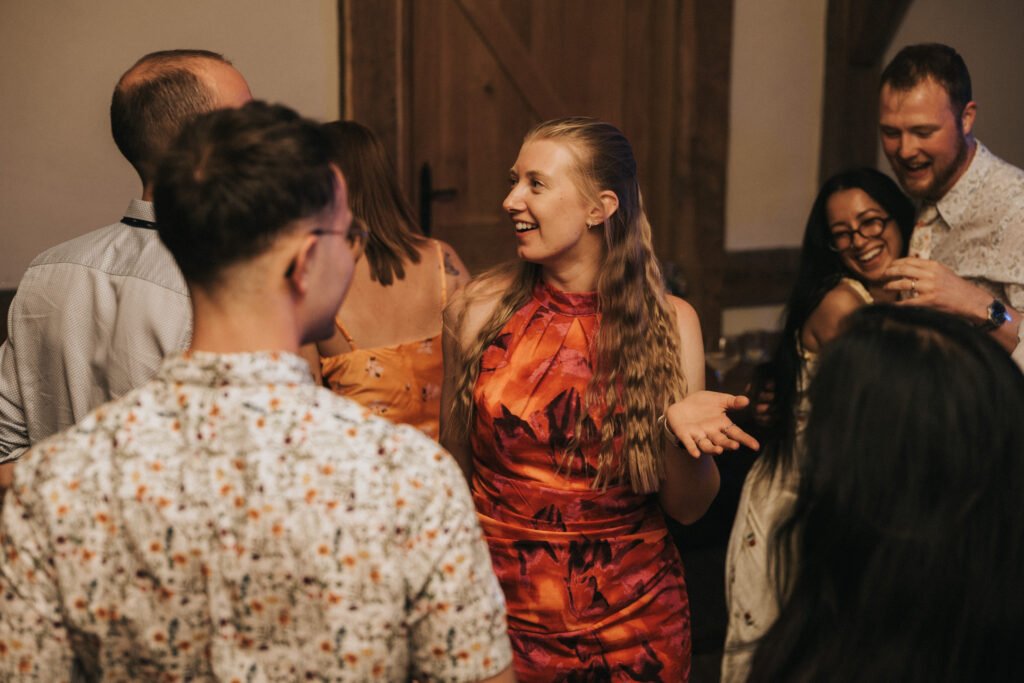 A woman in a red patterned dress smiles and gestures whilst talking with a group of people at a lively indoor social gathering. Others around her are chatting and laughing, creating a joyful atmosphere. © Aimee Lince Photography, Wedding photographer in Lincolnshire