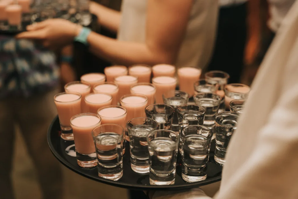 A person holds a tray filled with small glasses of clear liquid and pink creamy drinks, likely shots, at a social event. More people holding similar trays are visible in the background. © Aimee Lince Photography, Wedding photographer in Lincolnshire