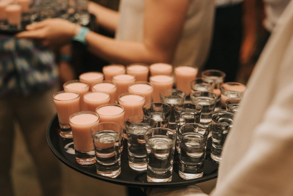 A person holds a tray filled with small glasses of clear liquid and pink creamy drinks, likely shots, at a social event. More people holding similar trays are visible in the background. © Aimee Lince Photography, Wedding photographer in Lincolnshire