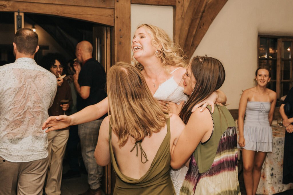 A joyful woman in a white dress laughs and hugs two friends on a dance floor at a lively indoor celebration, with other guests socialising in the background. © Aimee Lince Photography, Wedding photographer in Lincolnshire