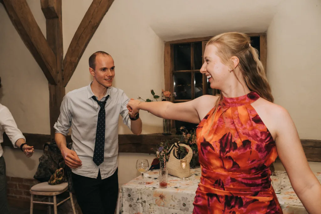 A smiling man in a shirt and loosened tie dances with a woman in a vibrant red and pink floral dress in a warmly lit room decorated with wooden beams and a table with flowers in the background. © Aimee Lince Photography, Wedding photographer in Lincolnshire