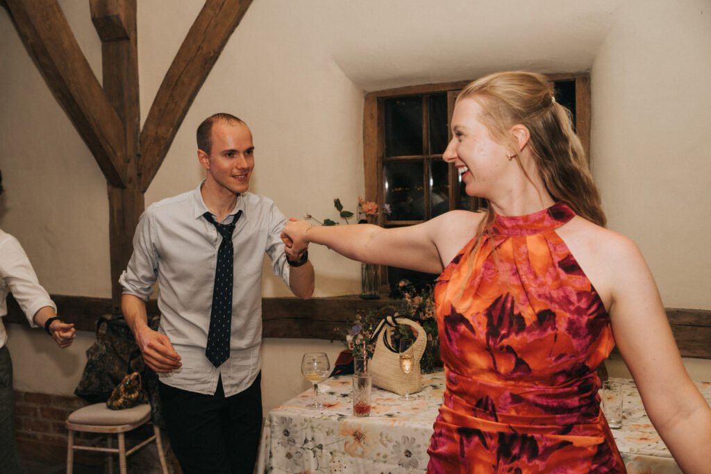 A smiling man in a shirt and loosened tie dances with a woman in a vibrant red and pink floral dress in a warmly lit room decorated with wooden beams and a table with flowers in the background. © Aimee Lince Photography, Wedding photographer in Lincolnshire