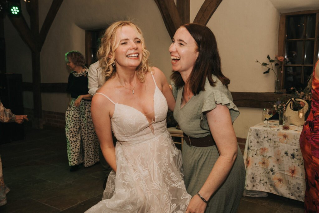 Two women joyfully laughing and dancing together at a celebration. One woman is wearing a white wedding dress, while the other is dressed in a light green dress. The setting appears festive with warmly lit décor and other people in the background. © Aimee Lince Photography, Wedding photographer in Lincolnshire
