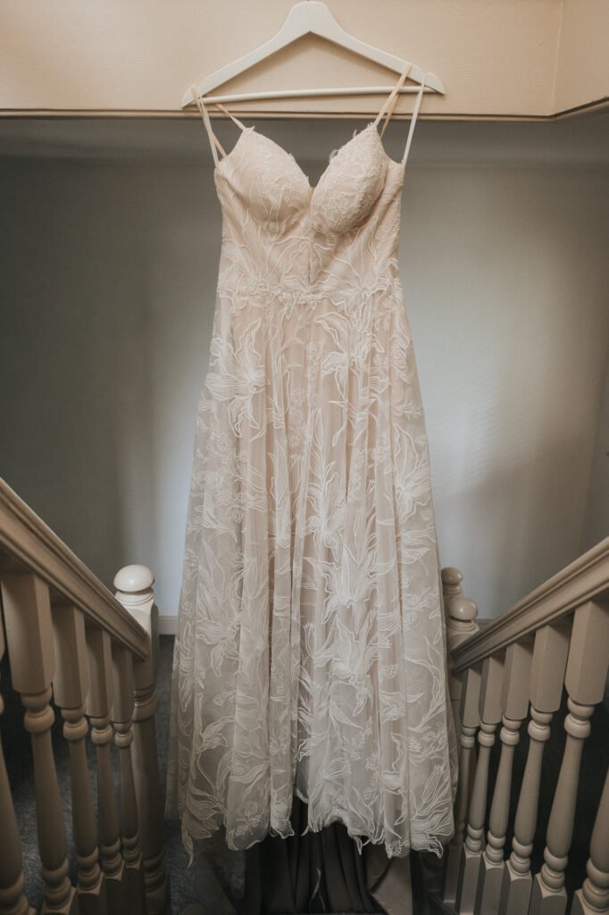 A white lace wedding dress with thin straps hangs on a white hanger above a staircase, displayed between beige banisters. The dress features a fitted bodice and a flowing, embroidered skirt. © Aimee Lince Photography, Wedding photographer in Lincolnshire