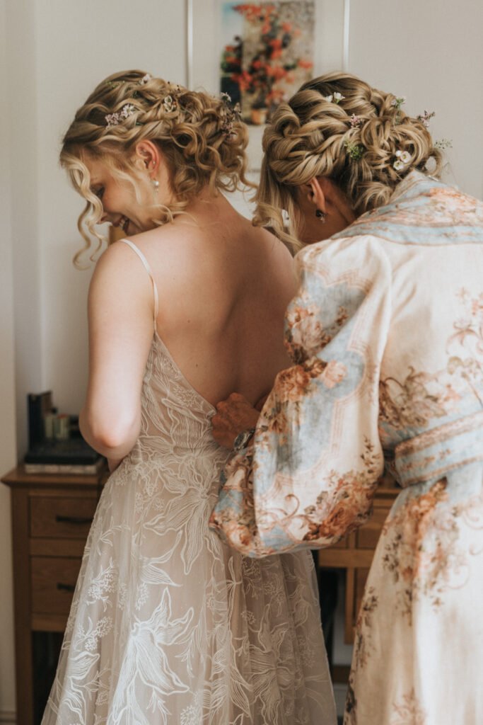A woman in a floral dressing gown helps do up the back of another woman’s delicate, lace wedding dress. Both have styled curly hair adorned with small flowers. They are in a softly lit room with wooden furniture and a floral picture on the wall. © Aimee Lince Photography, Wedding photographer in Lincolnshire