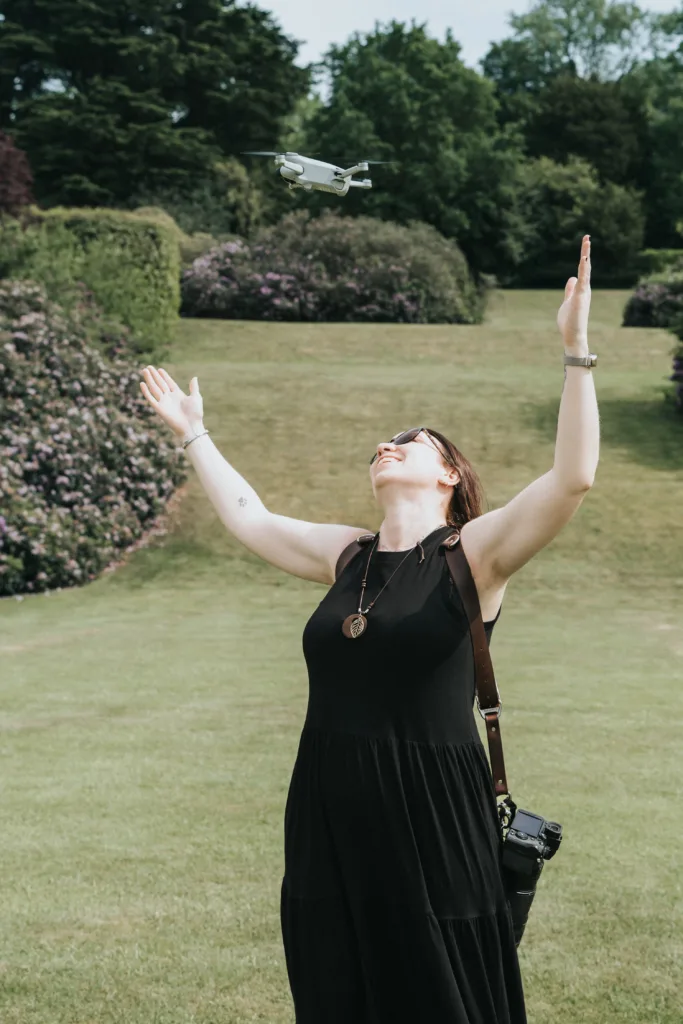 A woman in a black dress stands on a grassy lawn at Harlaxton Manor Wedding, arms raised and smiling up at a small drone. She wears sunglasses, a necklace, and carries a camera bag, surrounded by lush greenery and flowering shrubs on a sunny day. © Aimee Lince Photography, Lincolnshire photographer