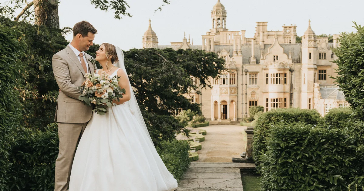 A bride in a white gown and veil holds a bouquet, smiling at her groom in a tan suit. They stand outdoors on a stone path framed by greenery, with the ornate, historic Harlaxton Manor in the background—a perfect wedding moment. © Aimee Lince Photography, Lincolnshire photographer