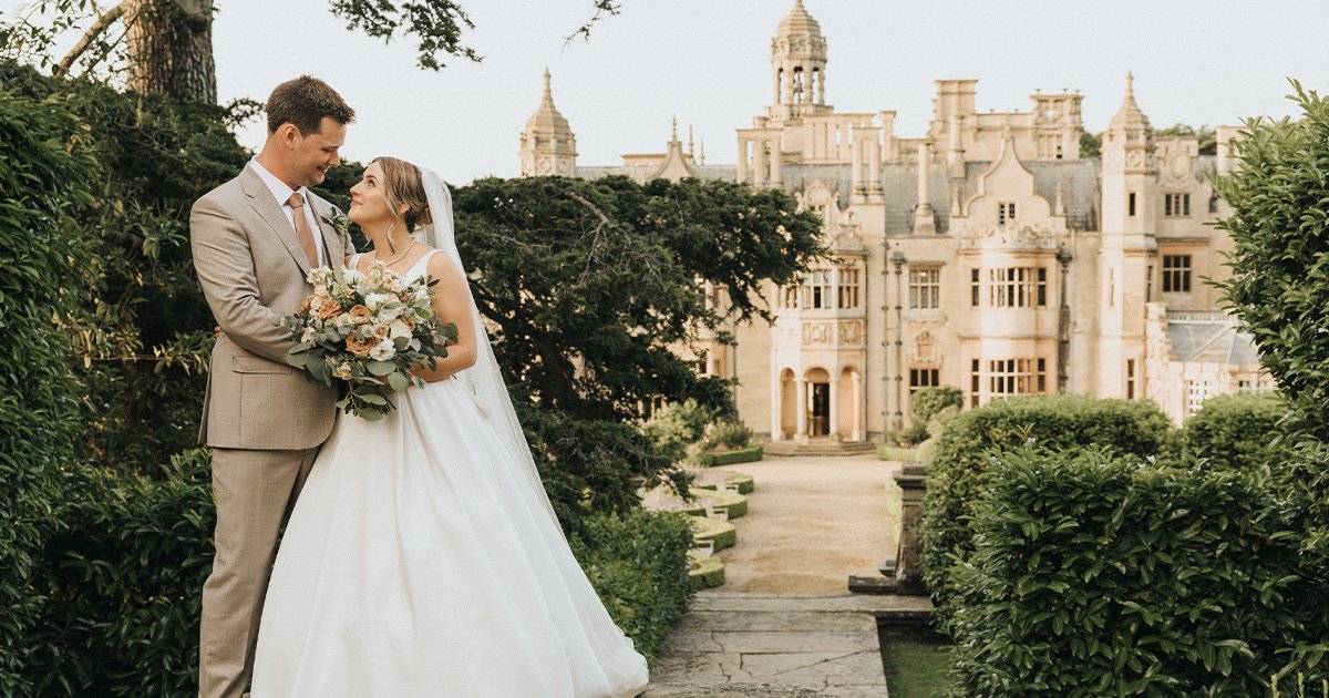 A bride in a white gown and veil holds a bouquet, smiling at her groom in a tan suit. They stand outdoors on a stone path framed by greenery, with the ornate, historic Harlaxton Manor in the background—a perfect wedding moment. © Aimee Lince Photography, Lincolnshire photographer