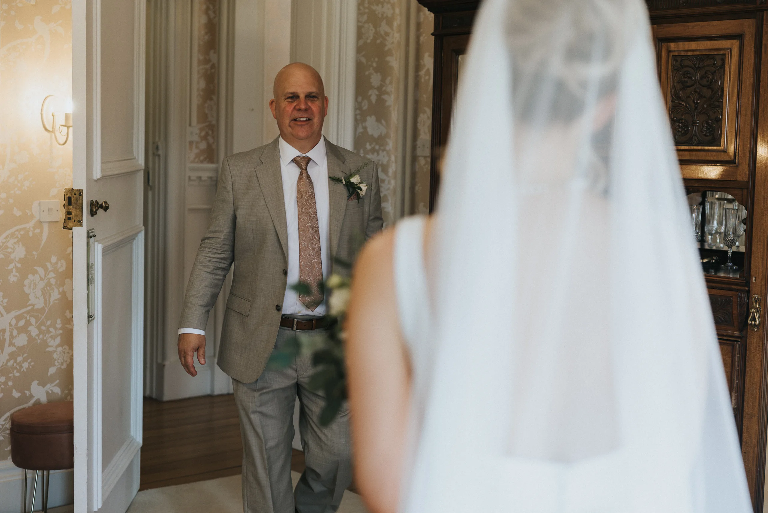 A smiling, bald man in a light grey suit and floral tie stands in a doorway at a Harlaxton Manor wedding, looking at a bride in a white dress and veil who faces him, slightly out of focus. The room features patterned wallpaper and wooden furniture. © Aimee Lince Photography, Lincolnshire photographer