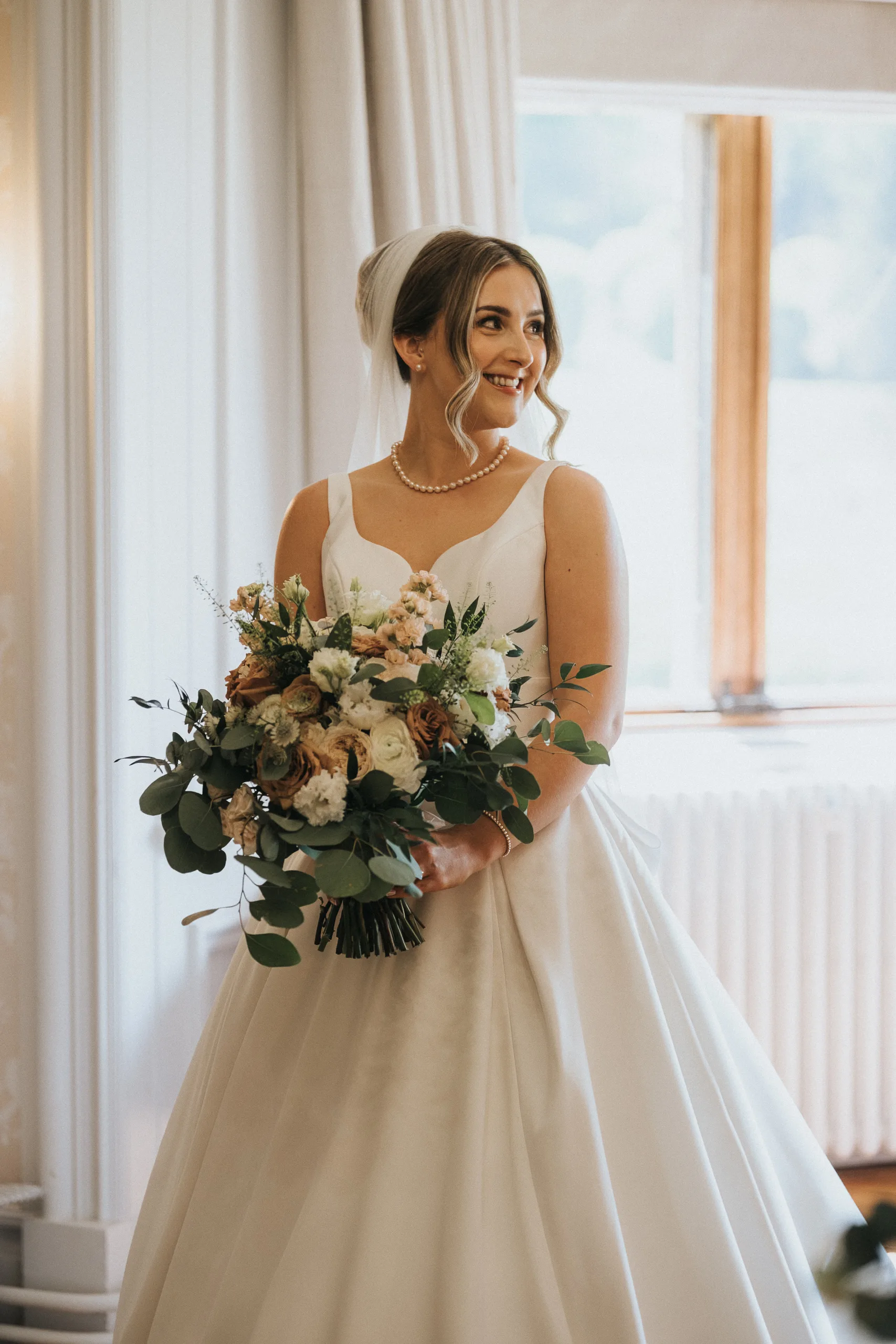 A bride in a white dress and veil stands indoors at her Harlaxton Manor Wedding, smiling to the side. She holds a large bouquet of greenery and muted pink blooms as light streams through the window, softly illuminating the scene. © Aimee Lince Photography, Lincolnshire photographer
