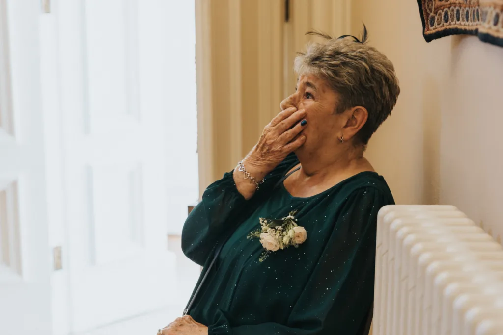 An elderly woman with short grey hair sits indoors at a Harlaxton Manor wedding, her hand covering her mouth in an emotional gesture. She wears a teal dress with a white corsage and silver bracelet, appearing moved as light streams through a nearby open door. © Aimee Lince Photography, Lincolnshire photographer