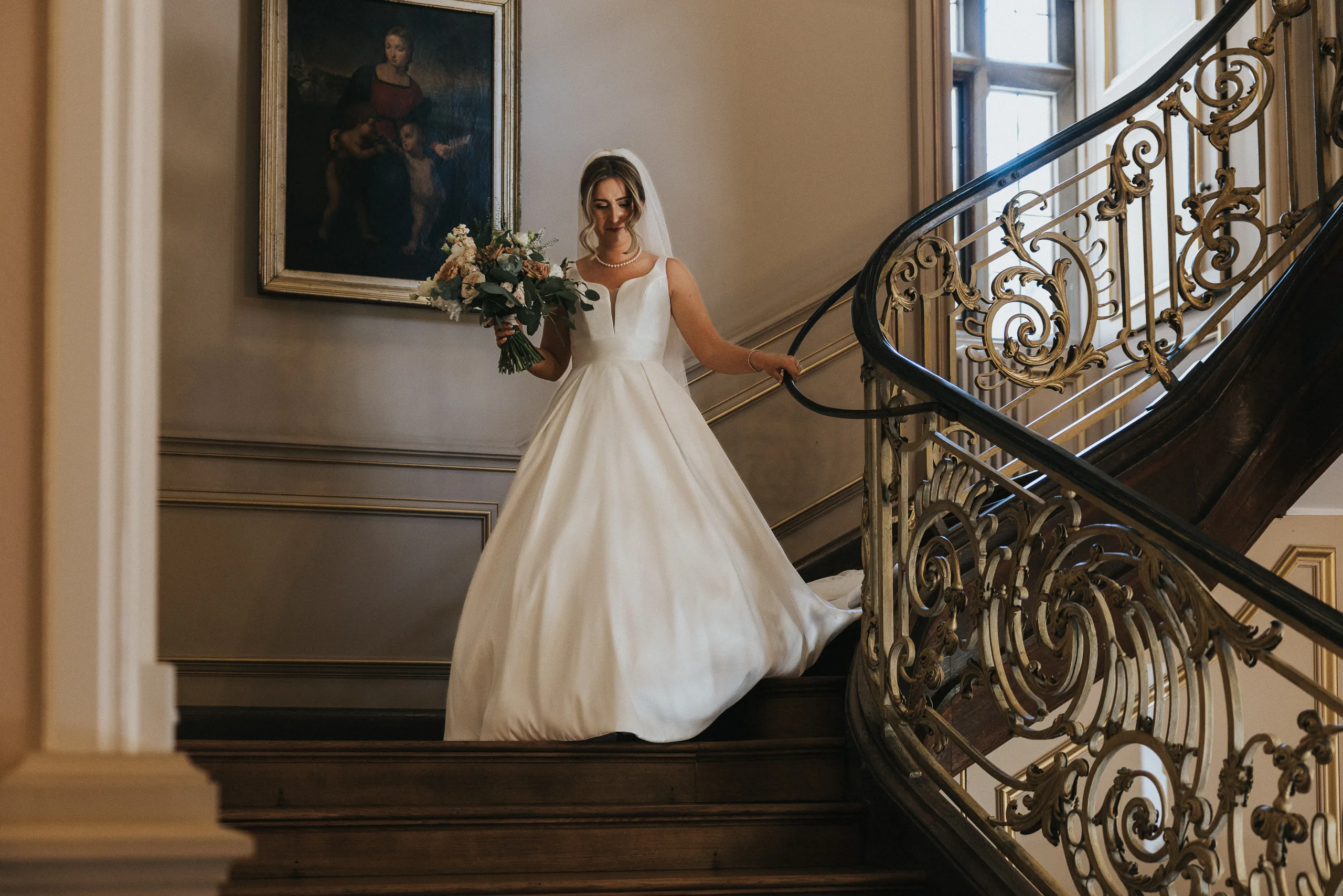 A bride in a white gown descends an ornate wooden staircase with decorative railings at her Harlaxton Manor Wedding, holding a bouquet. She wears a veil and smiles softly as sunlight streams through tall windows behind her. © Aimee Lince Photography, Lincolnshire photographer