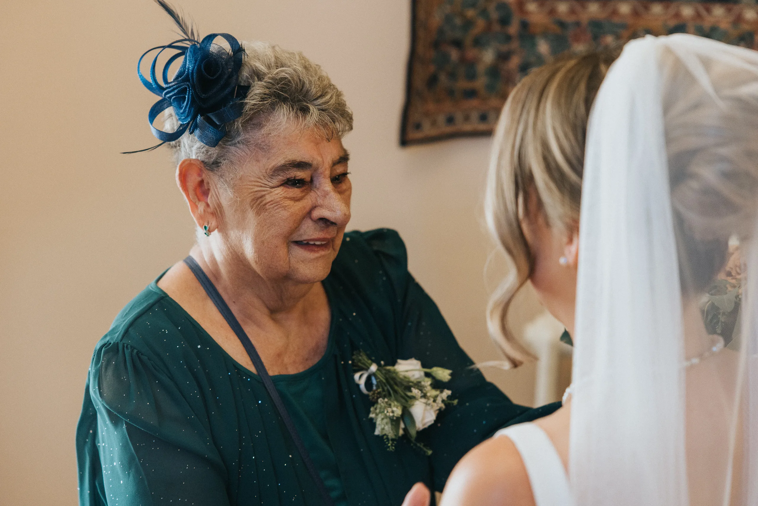 An elderly woman in a teal dress and navy fascinator smiles warmly at a bride in white. She wears a corsage and earrings; the bride’s back is to the camera. The warmly lit Harlaxton Manor Wedding setting features a tapestry in the background. © Aimee Lince Photography, Lincolnshire photographer