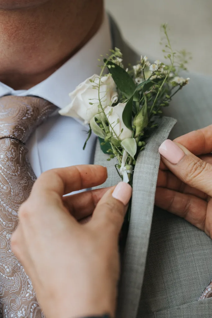 Close-up of hands pinning a white rose buttonhole with greenery onto the lapel of a light grey suit at a Harlaxton Manor Wedding. The person wears a paisley-patterned beige tie and white shirt. Pale pink varnish adorns their nails; the background is softly blurred. © Aimee Lince Photography, Lincolnshire photographer