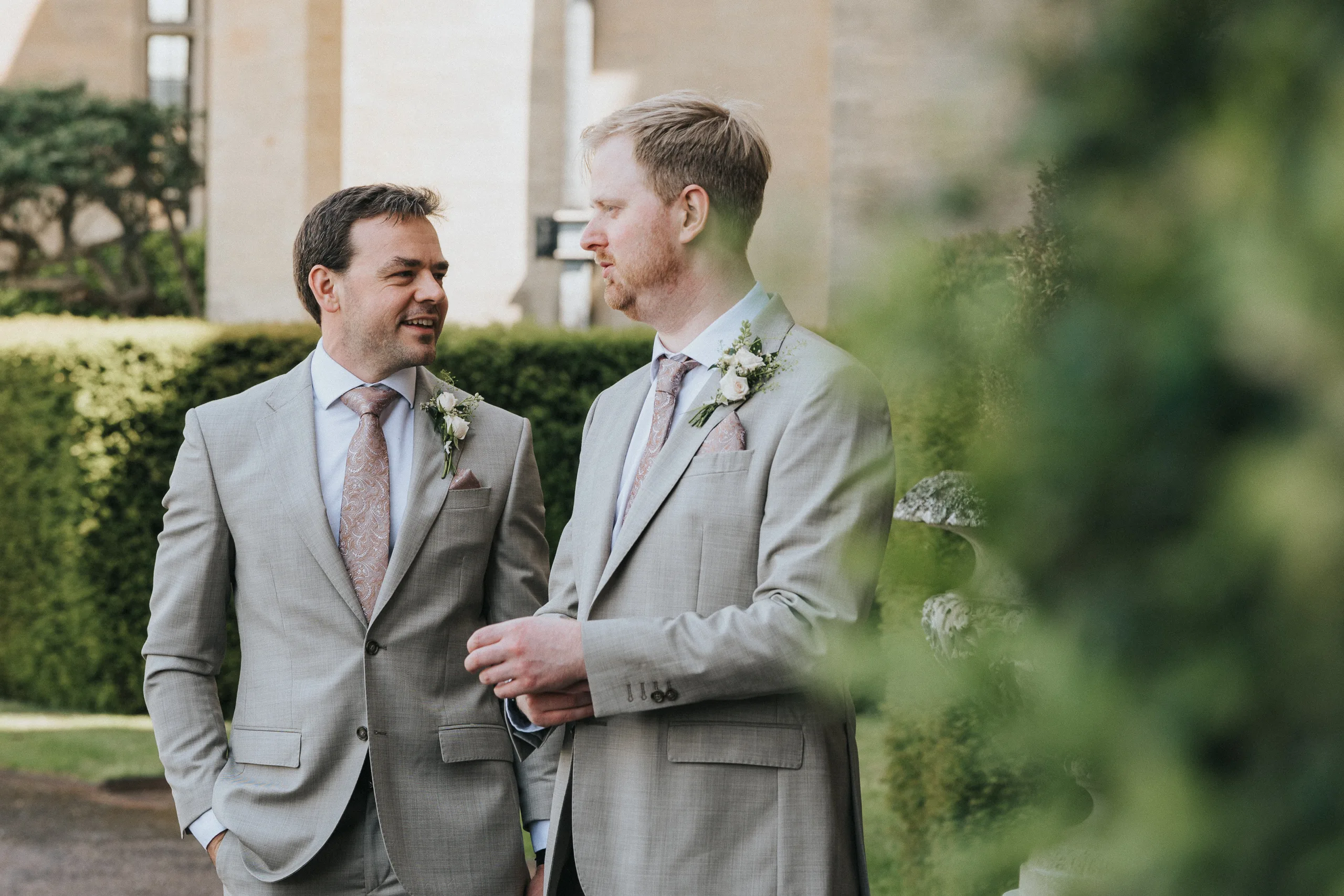 Two men in light grey suits with buttonholes and pink ties stand outdoors, talking and smiling at a Harlaxton Manor wedding. They are surrounded by greenery, with blurred bushes in the foreground and stone buildings behind. © Aimee Lince Photography, Lincolnshire photographer