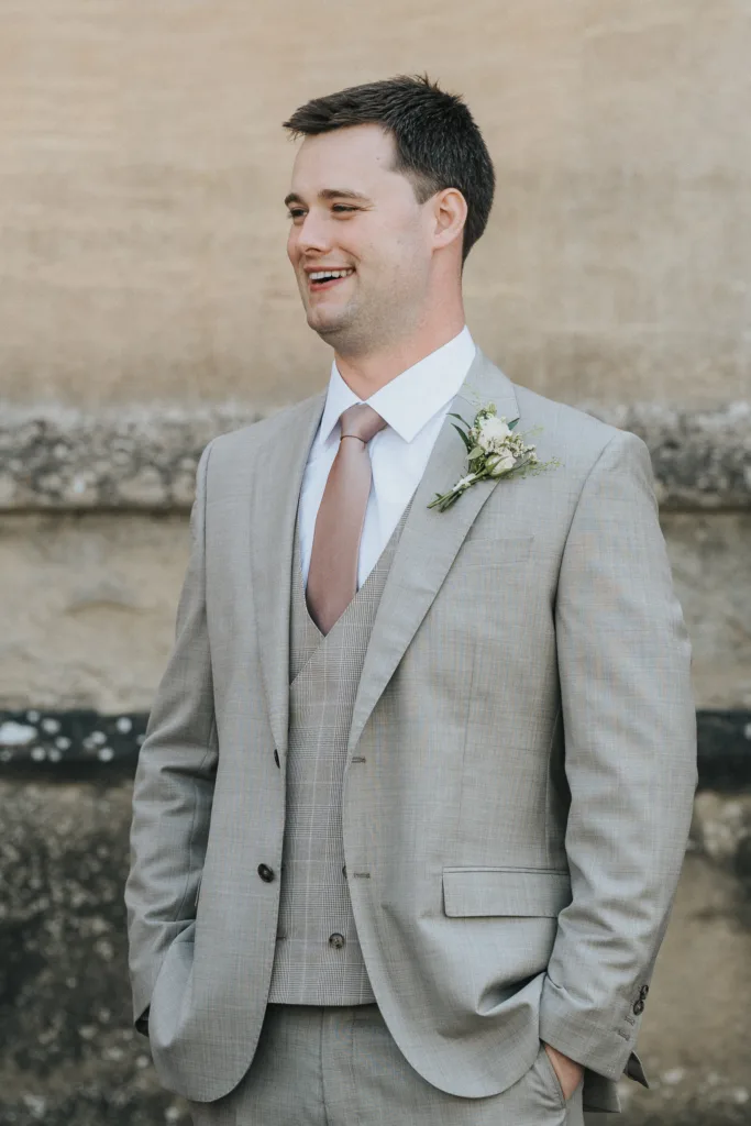 A man in a light grey suit with a matching waistcoat and light mauve tie stands smiling at his Harlaxton Manor wedding. He wears a white shirt and buttonhole with white flowers and greenery, standing before an elegant stone wall. © Aimee Lince Photography, Lincolnshire photographer