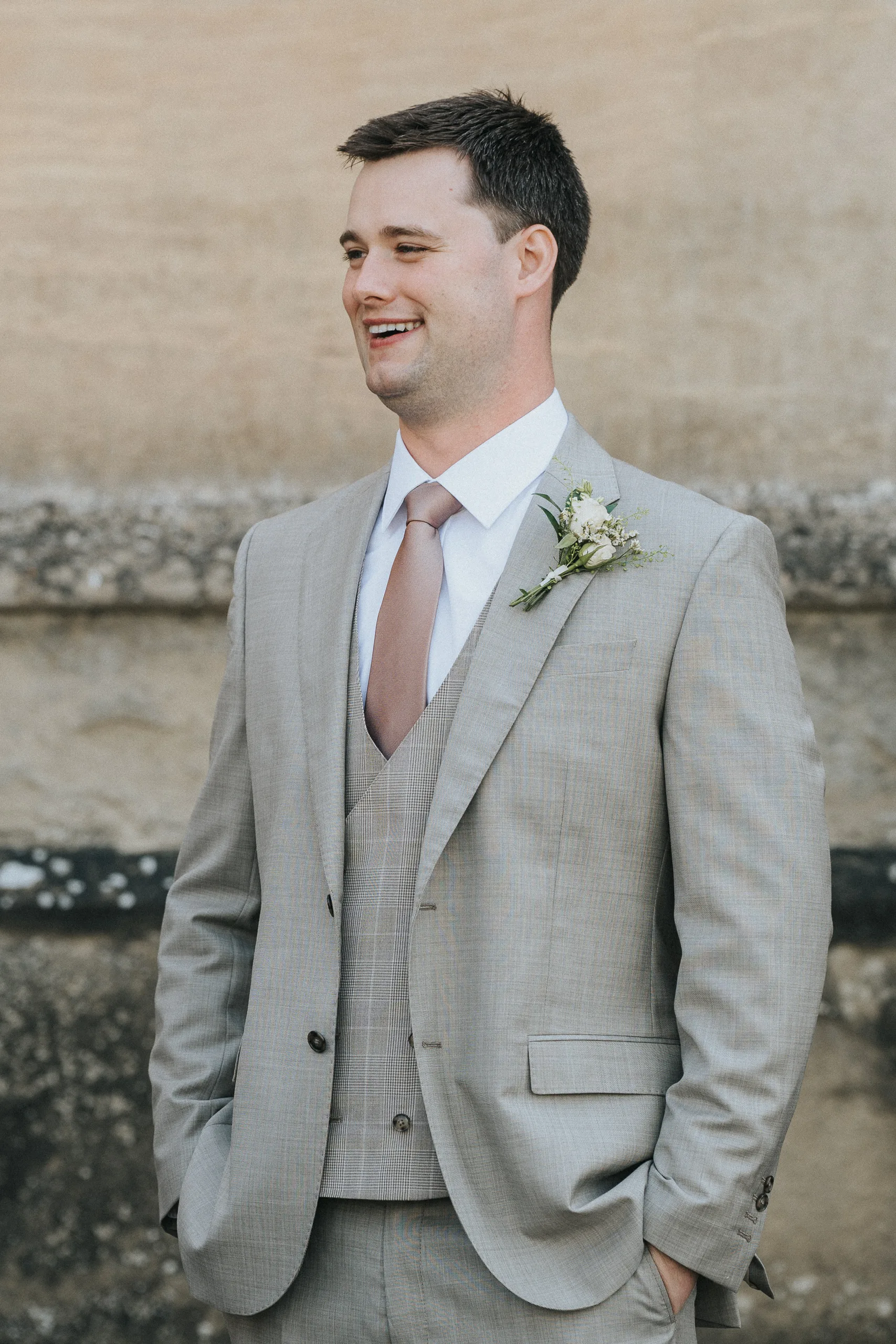 A man in a light grey suit with a matching waistcoat and light mauve tie stands smiling at his Harlaxton Manor wedding. He wears a white shirt and buttonhole with white flowers and greenery, standing before an elegant stone wall. © Aimee Lince Photography, Lincolnshire photographer
