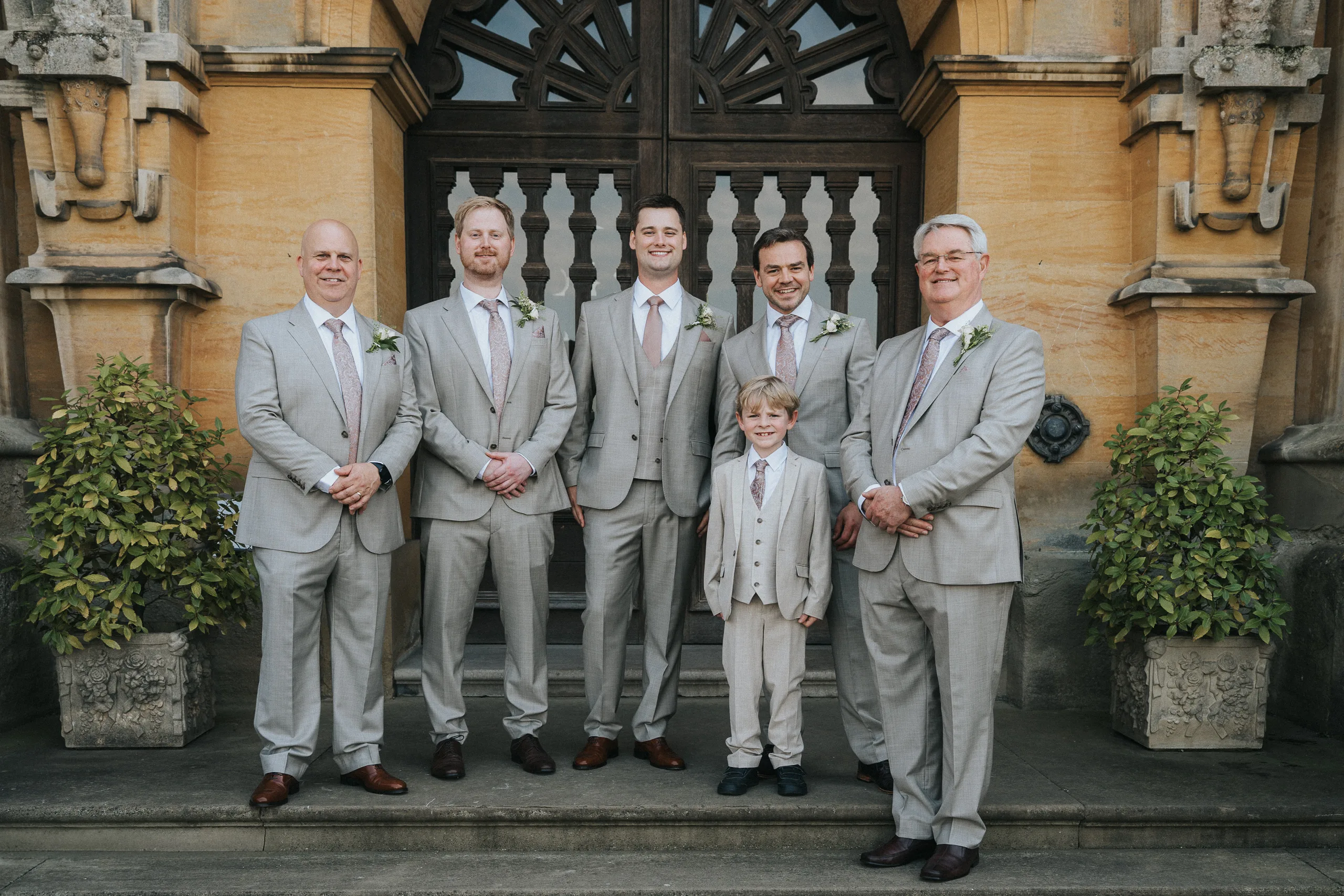Six men and a young boy in matching light grey suits with pale pink ties and buttonholes stand smiling on stone steps at a Harlaxton Manor Wedding, framed by potted plants, an ornate wooden door, and a grand yellow stone building. © Aimee Lince Photography, Lincolnshire photographer