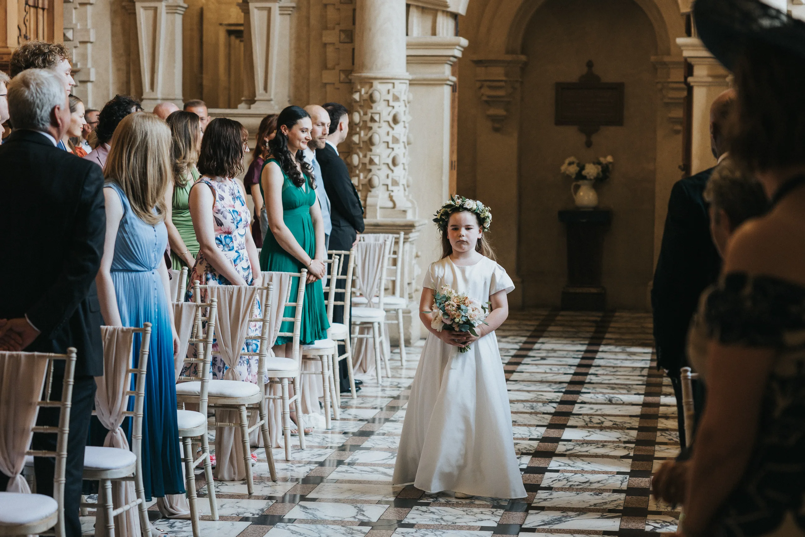 A young flower girl in a white dress and floral crown walks down the aisle at a Harlaxton Manor Wedding, surrounded by elegant columns, arches, and patterned tiled flooring that create a timeless, classical atmosphere. © Aimee Lince Photography, Lincolnshire photographer